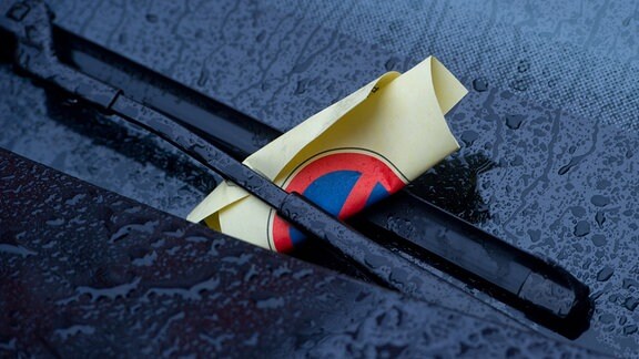 Symbolbild:  Ein Strafzettel (Knöllchen) steckt in einem Scheibenwischer eines geparkten Autos.