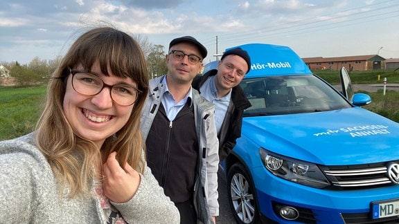 eine Frau und zwei Männer stehen vor einem blauen Auto