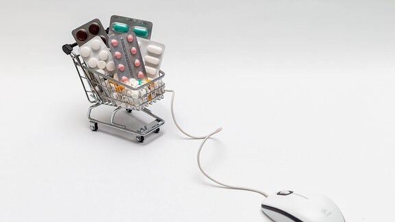 Eine Computermaus hängt an einem Mini-Einaufswagen mit Medikamenten drin.