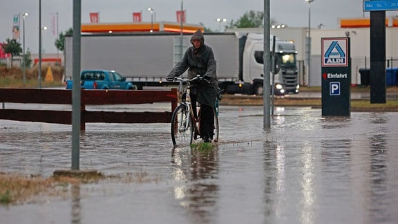 Eine Frau fährt nach einem Unwetter mit Starkregen mit ihrem Fahrrad über eine überflutete Straße in einem Gewerbegebiet.