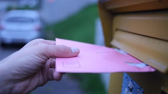 Eine Hand hält an einem Briefkasten einen Wahlbrief für die Stimmzettel der Wahlen für das Europaparlament, die Regionalversammlung und den Gemeinderat.