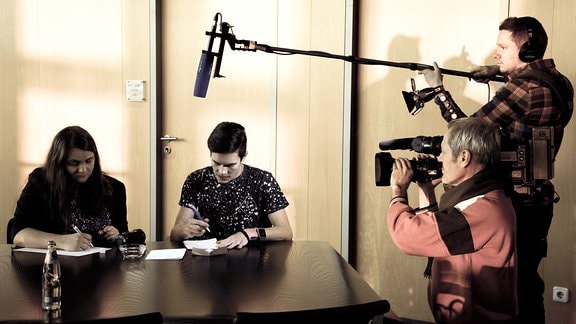 Zwei junge Erwachsene sitzen an einem Tisch und schreiben etwas auf Papier, sie werden von zwei weiteren Menschen per Videokamera gefilmt.