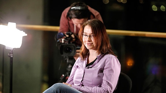 Eine junge Frau sitzt auf einem Stuhl, im Hintergrund ist ein Kameramann zu sehen.