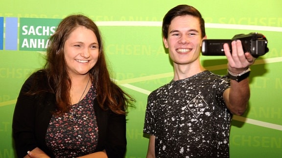 Zwei junge Erwachsene stehen lächelnd vor einer grünen Wand, der junge Mann hält eine Videokamera in die Hand.