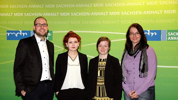 Vier Menschen stehen vor einer grünen Wand und lächeln in die Kamera.
