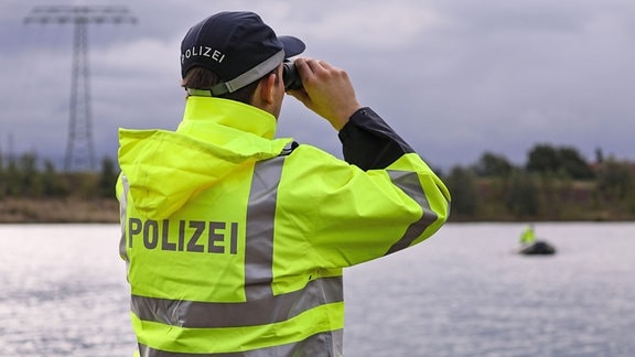 Ein Polizeibeamter in gelb-grüner Regenjacke mit der Aufschrift Polizei auf dem Rücken schaut mit dem Fernglas auf ein Gewässer.