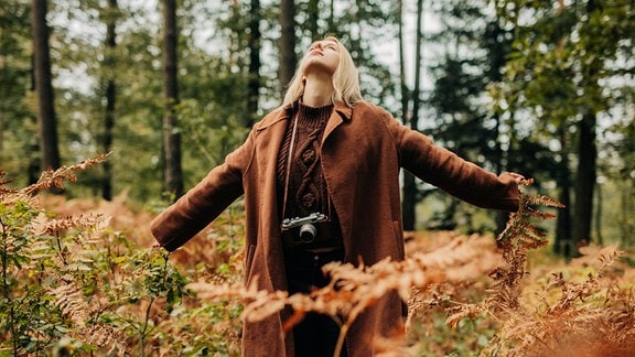 Eine blonde Frau in braunen Anziehsachen steht, die Arme ausgestreckt, in einer Herbstlandschaft.