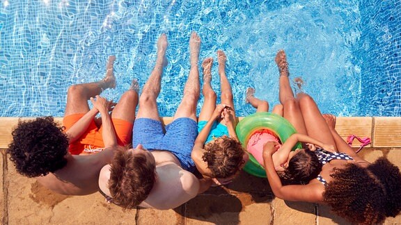 Familie sitzt in Schwimmbad am Beckenrand mit den Füßen im Wasser.