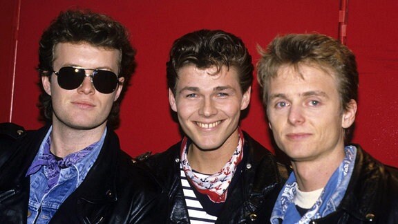Drei Männer posieren lächelnd für ein Foto vor einer roten Wand.