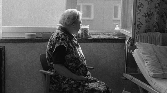 Eine alte Frau sitzt an einem geöffneten Fenster, gegenüber ist ein Plattenbau zu sehen.