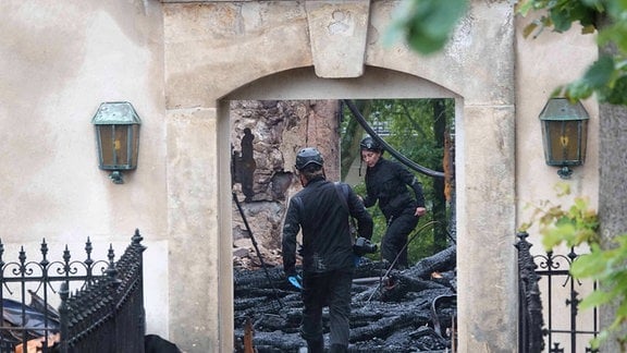 Eine Polizistin und ein Polizist laufen vorsichtig durch eine Ruine an verbrannten Holzbalken vorbei. Sie wurden am 8. August 2023 bei der Ermittlungsarbeit in der abgebrannten Kirche Großröhrsdorf fotografiert.