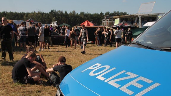 Leute feiern ein Festival im freien Gelände, ein Polizeiauto steht im Vordergrund.