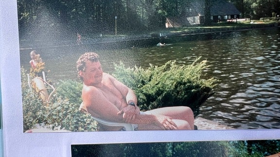 Ein abfotografiertes Foto von einem Mann in Badehosen auf einem Stuhl an einem See.