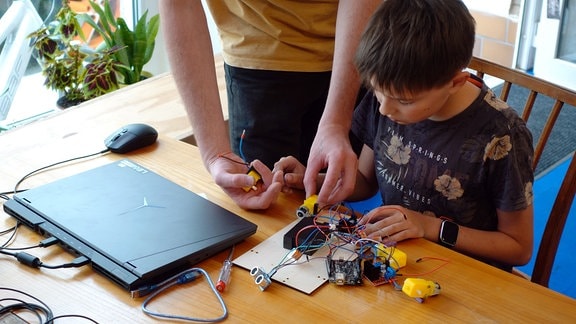 Ein Kind sitzt vor Elektrotechnik. Ein zugeklappter Laptop liegt danaben. Hände eines Erwachsenen zeigen etwas an den Kabeln. 