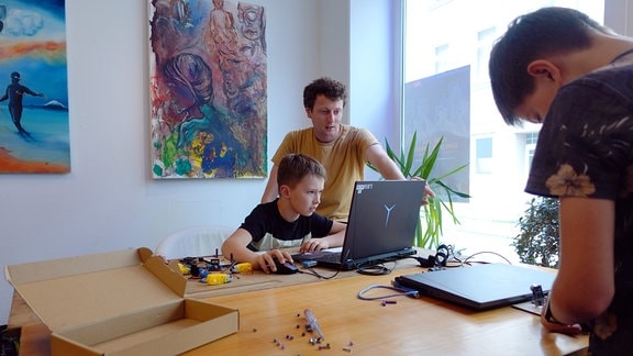 Ein Junge sitzt vor einem Laptop. Neben ihm ein erwachsener Mann, der etwas auf dem Bildschirm zeigt. 