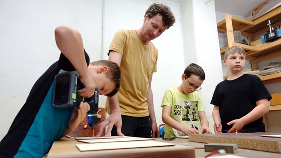 Drei Kinder und ein Mann stehen in einer Werkstatt. Der Mann hält ein Brett auf dem Tisch fest, was eines der Kinder bohrt.