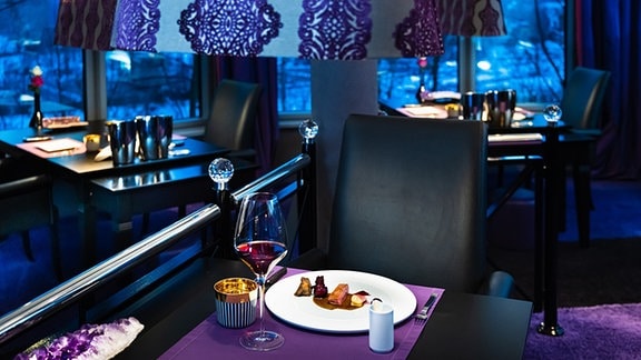 Ein Restaurant wurde in dunklen Materialien edel ausgestattet. Auf einem weiß gedeckten Tisch stehen Teller und Gläser.