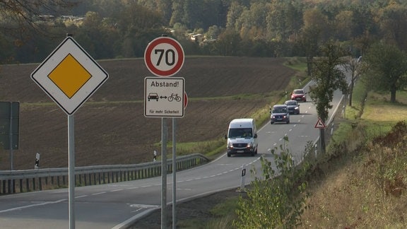 Eine Reihe Autos fahren auf einer Landstraße. Im Vordergrund stehen diverse Verkehrsschilder.