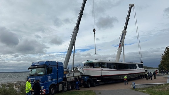 Ein Fahrgastschiff auf einem Riesenlaster wird von 2 Kränen heruntergehievt.