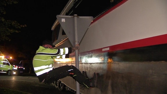 Ein Mann biegt mit aller Kraft ein Straßenverkehrsschild zur Seite, damit ein großes Schiff auf einem riesigen Laster nachts über die Straßen vorbei kommt.