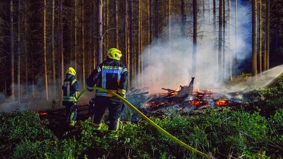 Mehrere Feuerwehrleute in Uniformen stehen vor einem Feuer im Wald und löschen die Flammen.