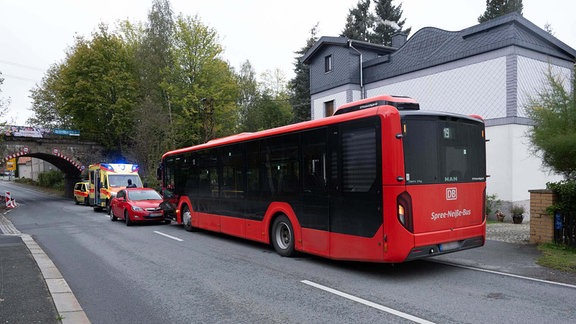 Ein langer roter Buns und ein roter Kleinwagen sowie Rettungsfahrzeuge stehen vor einer Bahnunterführung. 