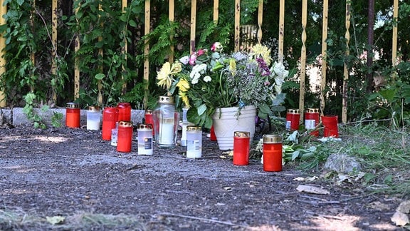 In der Ecke eines Parkplatzes in Schönau-Berzdorf auf dem Eigen stehen und liegen Blumensträuße und Grablichter.