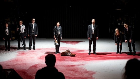Sieben Menschen in schwarzen Anzügen stehen in einer Reihe auf einer weißen Bühne mit einem riesigen roten Fleck.