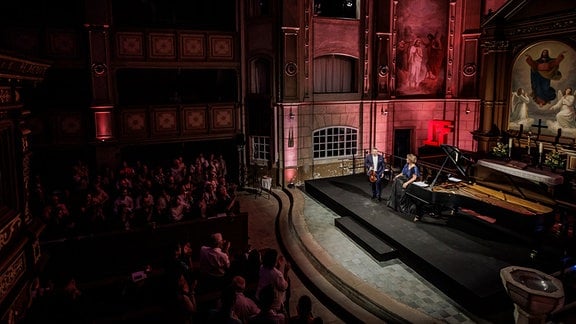 Zwei Musiker geben ein Konzert auf einer Bühne in einer Kirche, das Publikum applaudiert.