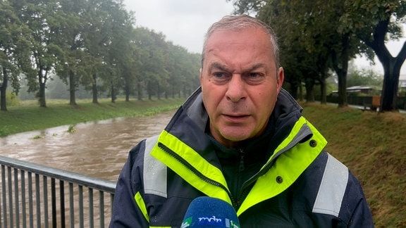 Ein Mann in einen blau-gelben Jacke steht an einem Fluss (Hochwasser) und gibt ein Interview.