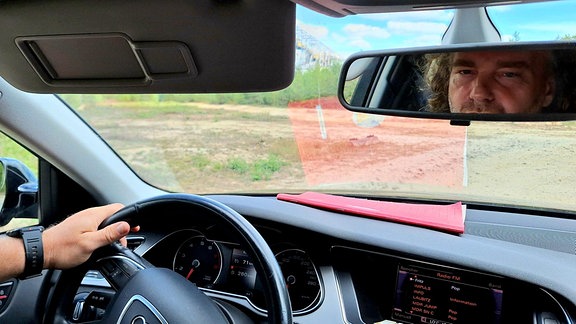 ein mann sitzt am Steuer seines Autos und blickt in den Rückspiegel. Dort sieht man sein Gesicht.