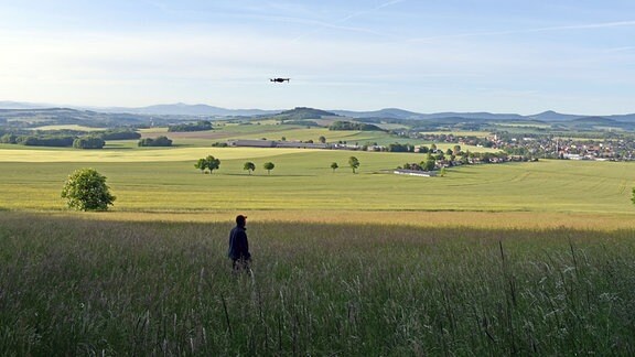 Eine Drohne fliegt über einer Wiesenlandschaft, im Vordergrund steht ein Mann.