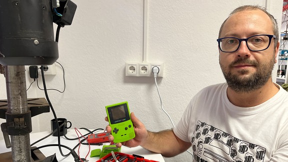 Ein Mann sitzt an einem Tisch neben einer Bohrmaschine und hält einen Gameboy in der Hand