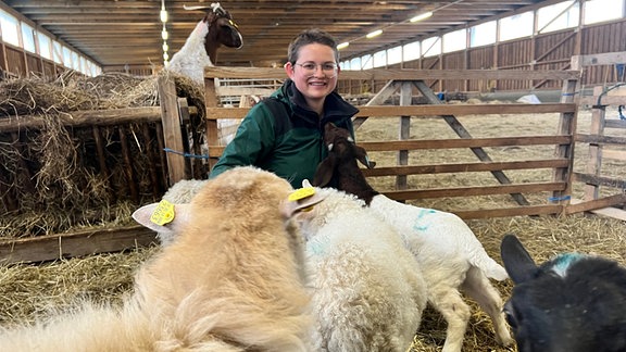 Eine junge Frau sitzt, umgeben von Schafen, in einem Stall