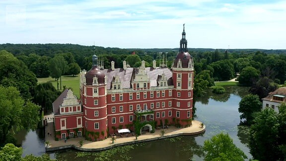 Blick auf das rote Schloss in Bad Muskau, umgeben von einem See und grünen Bäumen.
