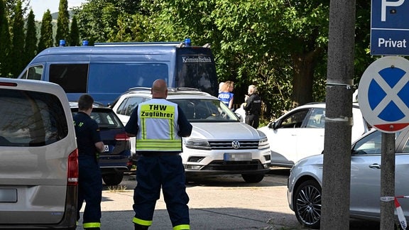 Auf einem Parkplatz stehen Autos, ein Fahrzeug mit der Aufschrift "Kriminaltechnik" sowie Polizisten und Mitarbeiter des Technischen Hilfswerks.