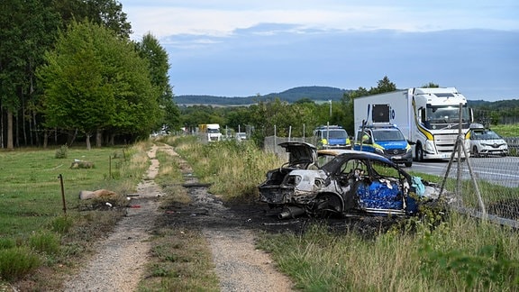 Neben einer teilweise gesperrten Autobahn liegt ein vollständig ausgebranntes Fahrzeug quer zur Fahrtrichtung.