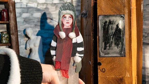 Eine Papierpuppe, die ein Mädchen mit Mütze und Schal zeigt, wird von einer Hand gehalten.