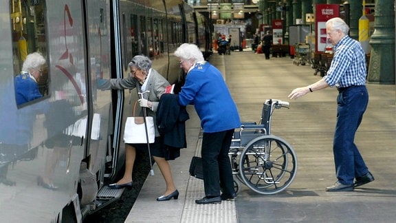 Eine ältere Frau steigt aus ihrem Rollstuhl in eine Bahn, eine Frau hilft ihr dabei.