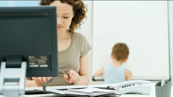 Ein Frau arbeitet an einem Computer und im Hintergrund ist ein Kind.