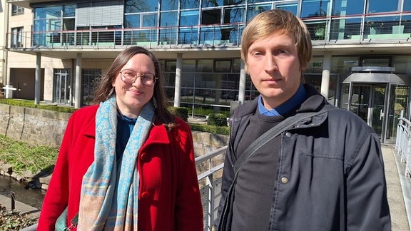 Eine Frau Anfang 40 steht rechts neben einem jüngeren Mann auf einem Steg zu einem gläsernen Gebäude. Die beiden sind Mandantin und Anwalt auf dem weg ins Verwaltungsgericht Chemnitz.