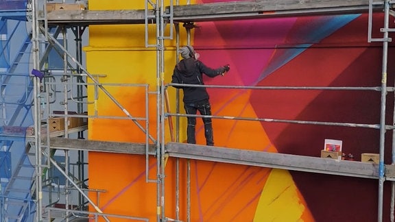 Eine Künstlerin auf einem Gerüst an einer bunten Wand mit Spraydosen in der Hand.