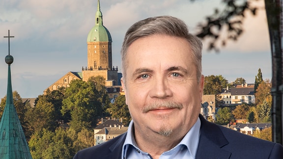 Rolf Schmidt, Oberbürgermeister von Annaberg-Buchholz, schaut in die Kamera, im Hintergrund eine Aufnahme der Stadt.