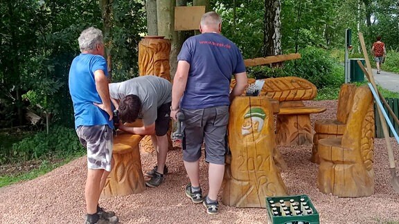 Menschen stehen in der Natur neben aus Holz geschnitzten Sitzen.