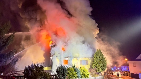 Ein Haus steht in Rauch und Flammen. Feuerwehrleute löschen.
