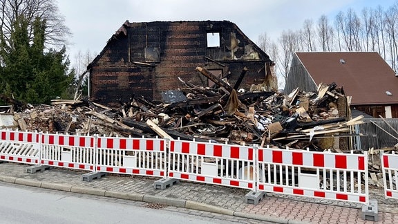 Ein abgebranntes Haus mit Trümmerteilen, davor der Fußweg mit Absperrzaun