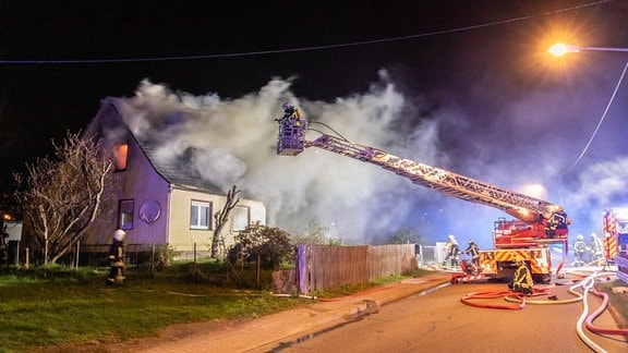 Aus einem Haus schlagen Flammen, Rauch steigt auf. Vor dem Haus steht ein Feuerwehrauto. Die Drehleiter ist ausgefahren. Ein Feuerwehrmann steht auf der Leiter und löscht das Feuer. 