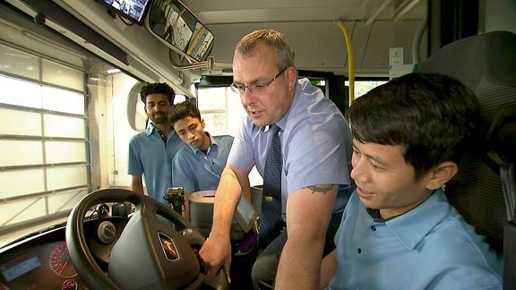 Ein Mann zeigt drei jungen Männern etwas am Steuerrad eines Busses.