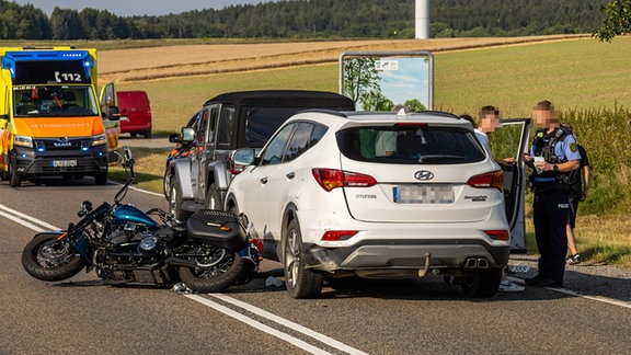 Eine Unfallstelle mit beschädigten Autos und einem Motorrad auf einer Landstraße. 