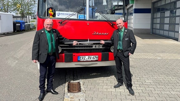 Zwei Busfahrer stehen vor einem roten Ikarus-Bus und halten ein Logo eines Motorenherstellers.